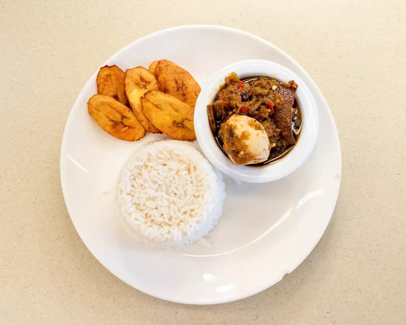 A plate of white rice, fried plantains, and a stew with meat and a hard-boiled egg.