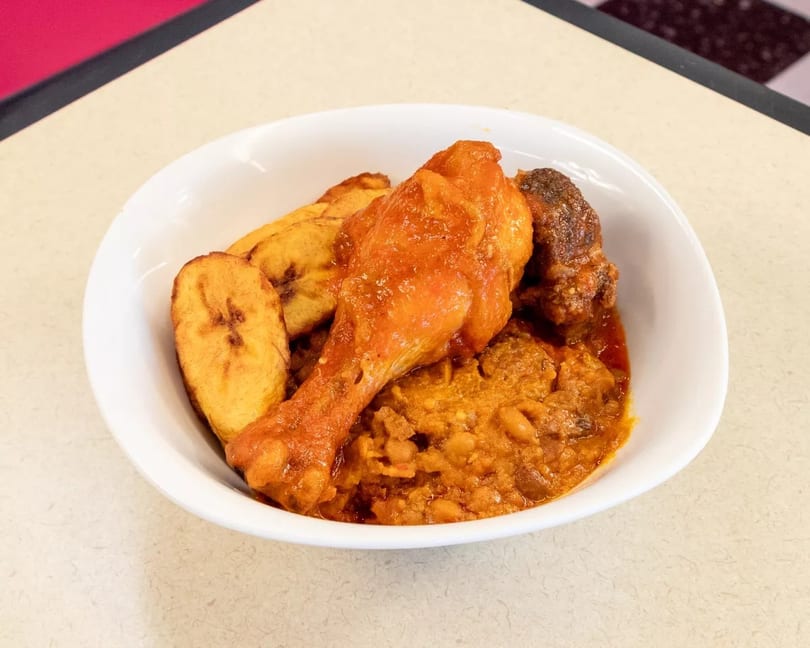 A white bowl filled with chicken, plantains, and a rich stew on a light-colored table.