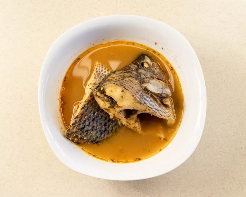 A white bowl filled with a rich orange broth and a whole cooked fish head and body.