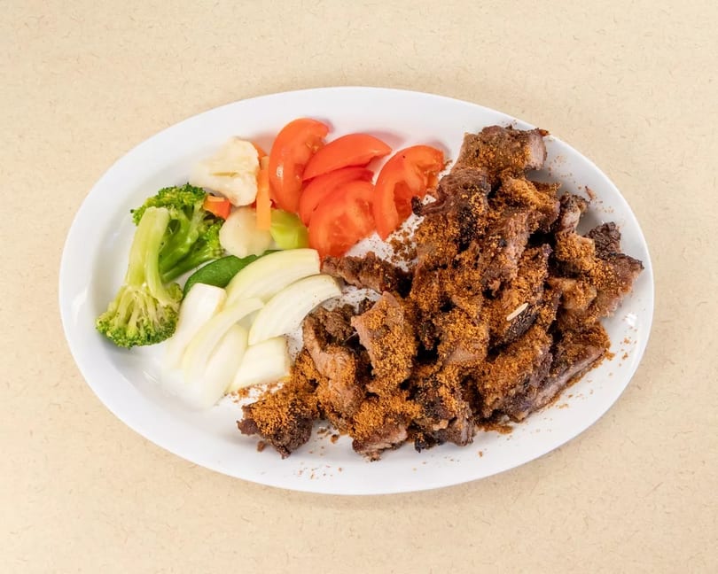Grilled meat with seasoning, sliced tomatoes, onions, broccoli, and cauliflower on a white plate.