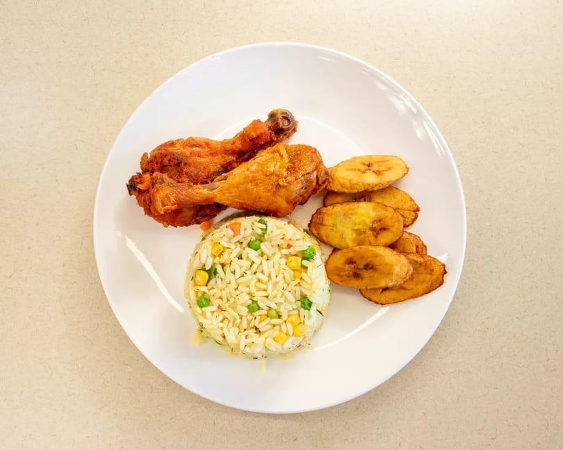 Fried chicken drumsticks, vegetable rice, and plantain slices on a white plate.